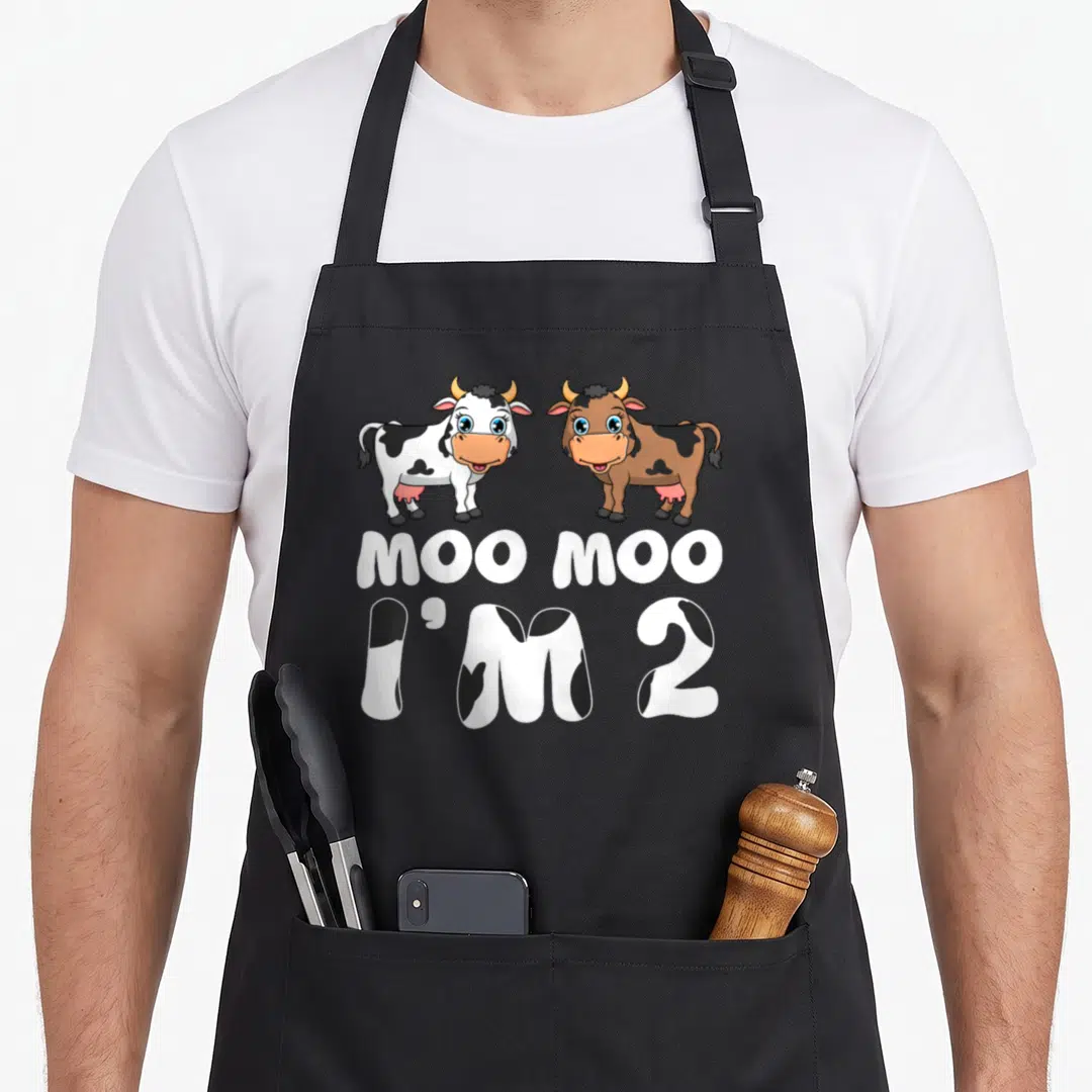 2nd cow apron - Two cartoon cows above MOO MOO I'M 2 text with cow spot pattern fill — shown on a black cotton canvas apron against a white background