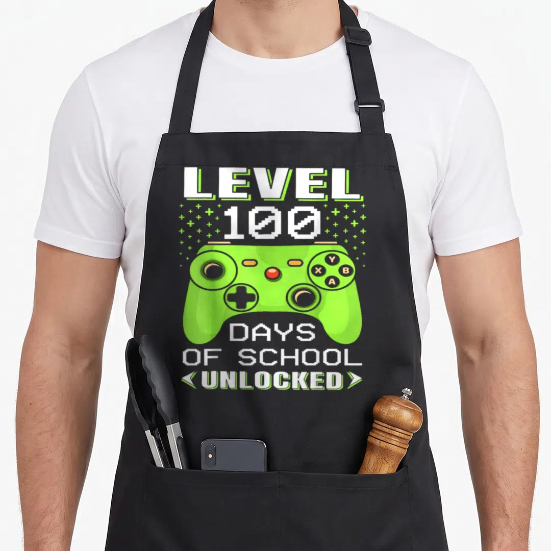 Video teacher apron - Lime green game controller with LEVEL 100 DAYS OF SCHOOL UNLOCKED text and plus signs — shown on a black cotton canvas apron against a white background