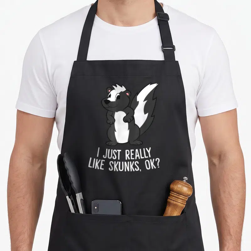 Skunk novelty apron - Cartoon skunk with white text reading I Just Really Like Skunks Ok — shown on a black cotton canvas apron against a white background