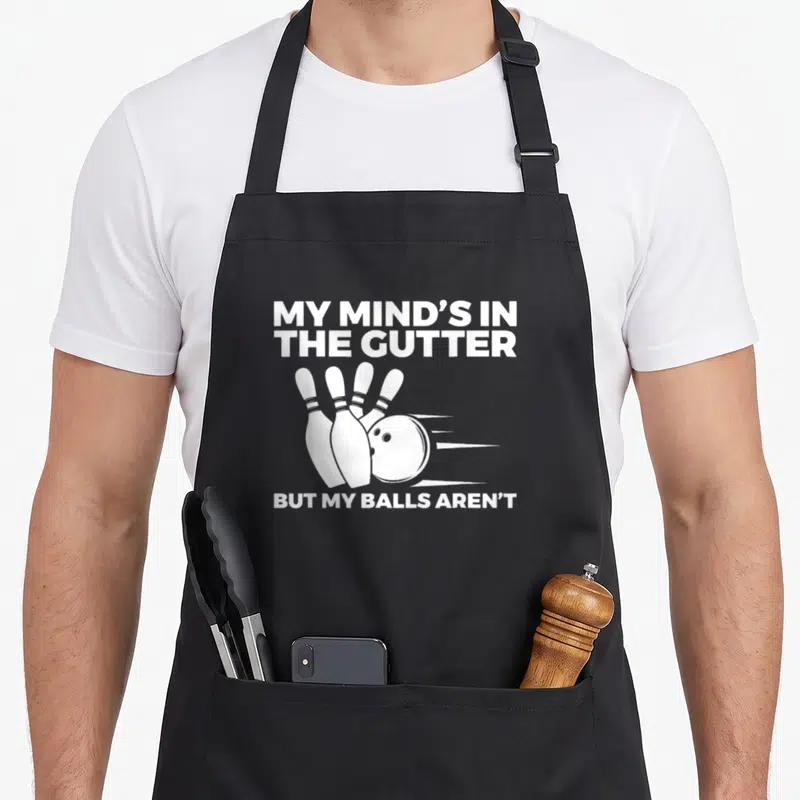 Bowling gutter apron - White text MY MIND'S IN THE GUTTER above a bowling ball striking four pins — shown on a black cotton canvas apron against a white background