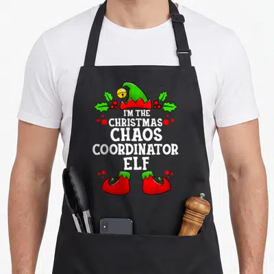 Chao christmas elf apron - I'm the Christmas Chaos Coordinator Elf text with green hat, holly, and red shoes — shown on a black cotton canvas apron against a white background