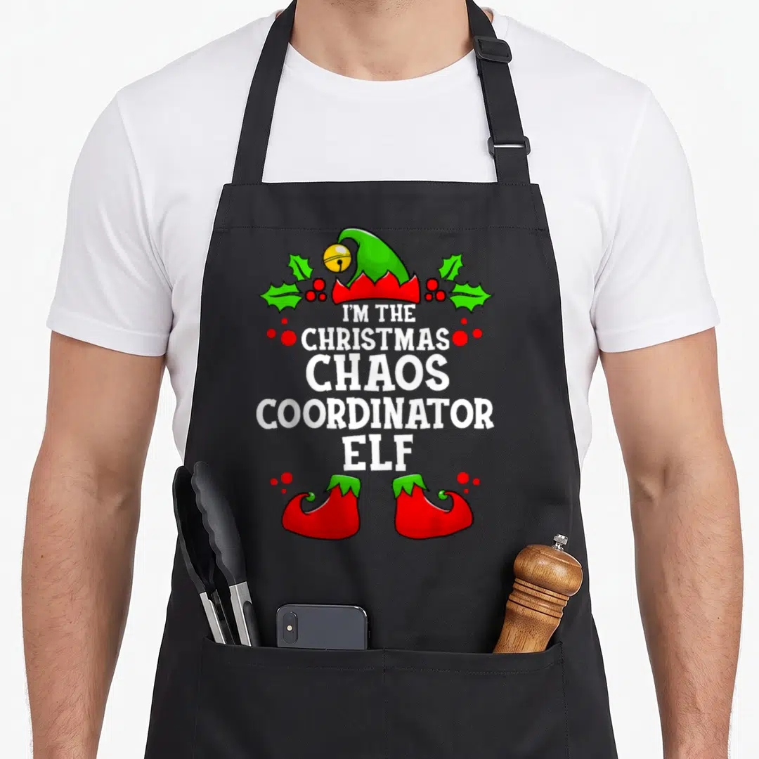 Chao christmas elf apron - I'm the Christmas Chaos Coordinator Elf text with green hat, holly, and red shoes — shown on a black cotton canvas apron against a white background