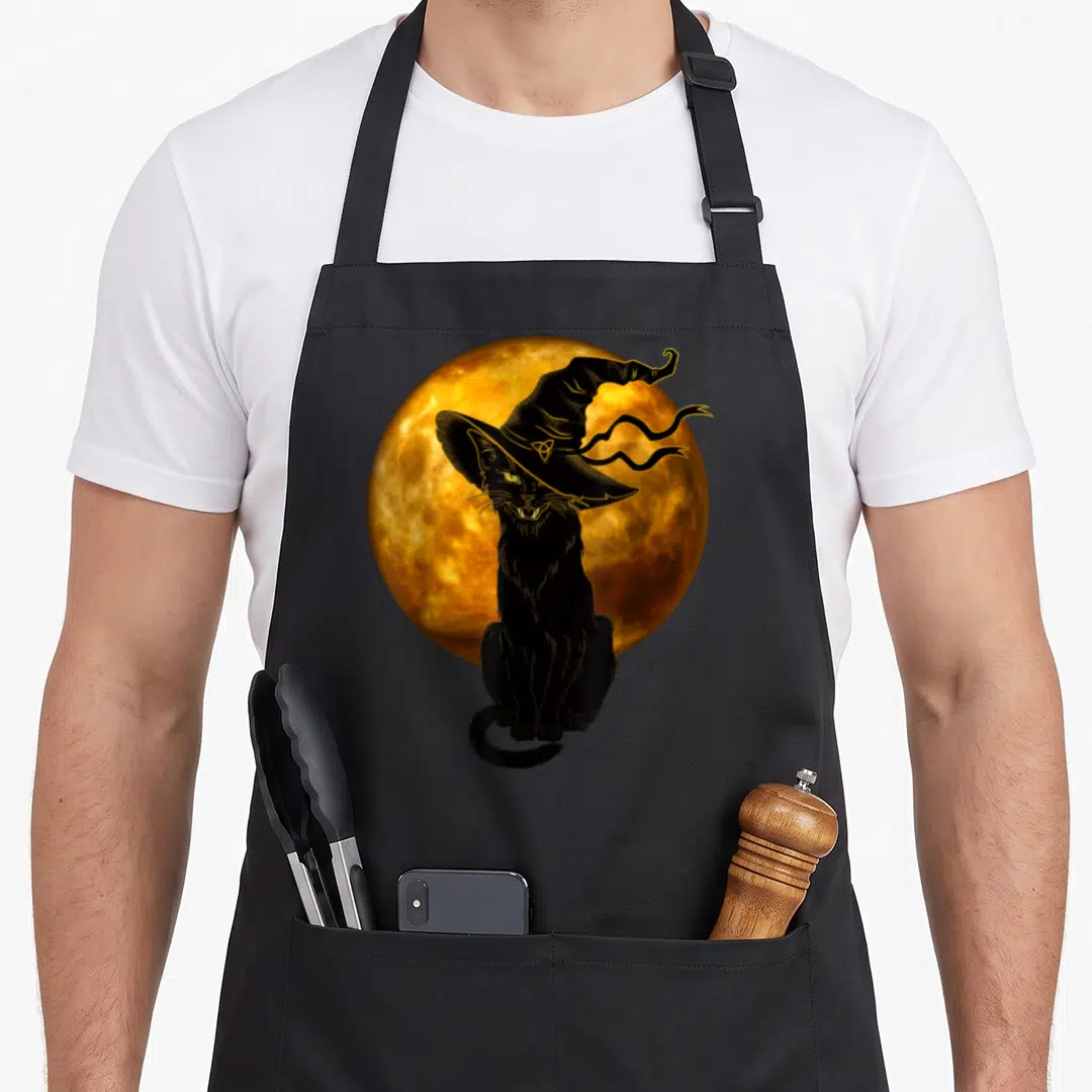 Scary halloween witch apron - Black cat wearing witch hat seated before orange full moon with gold outline details — shown on a black cotton canvas apron against a white background
