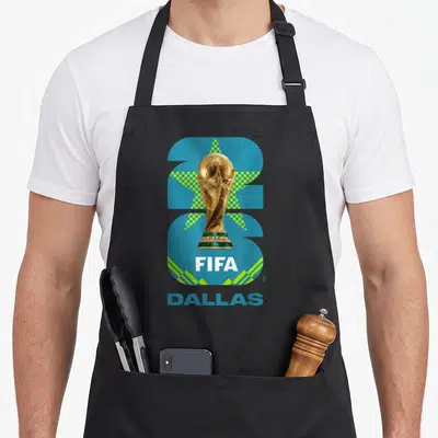 Cup26 trophy apron - FIFA World Cup trophy centered on blue shape with DALLAS text below — shown on a black cotton canvas apron against a white background