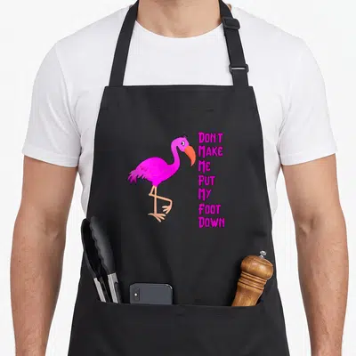 Retro awareness flamingo apron - Cartoon pink flamingo with orange beak next to stacked magenta text reading DON'T MAKE — shown on a black cotton canvas apron against a white background