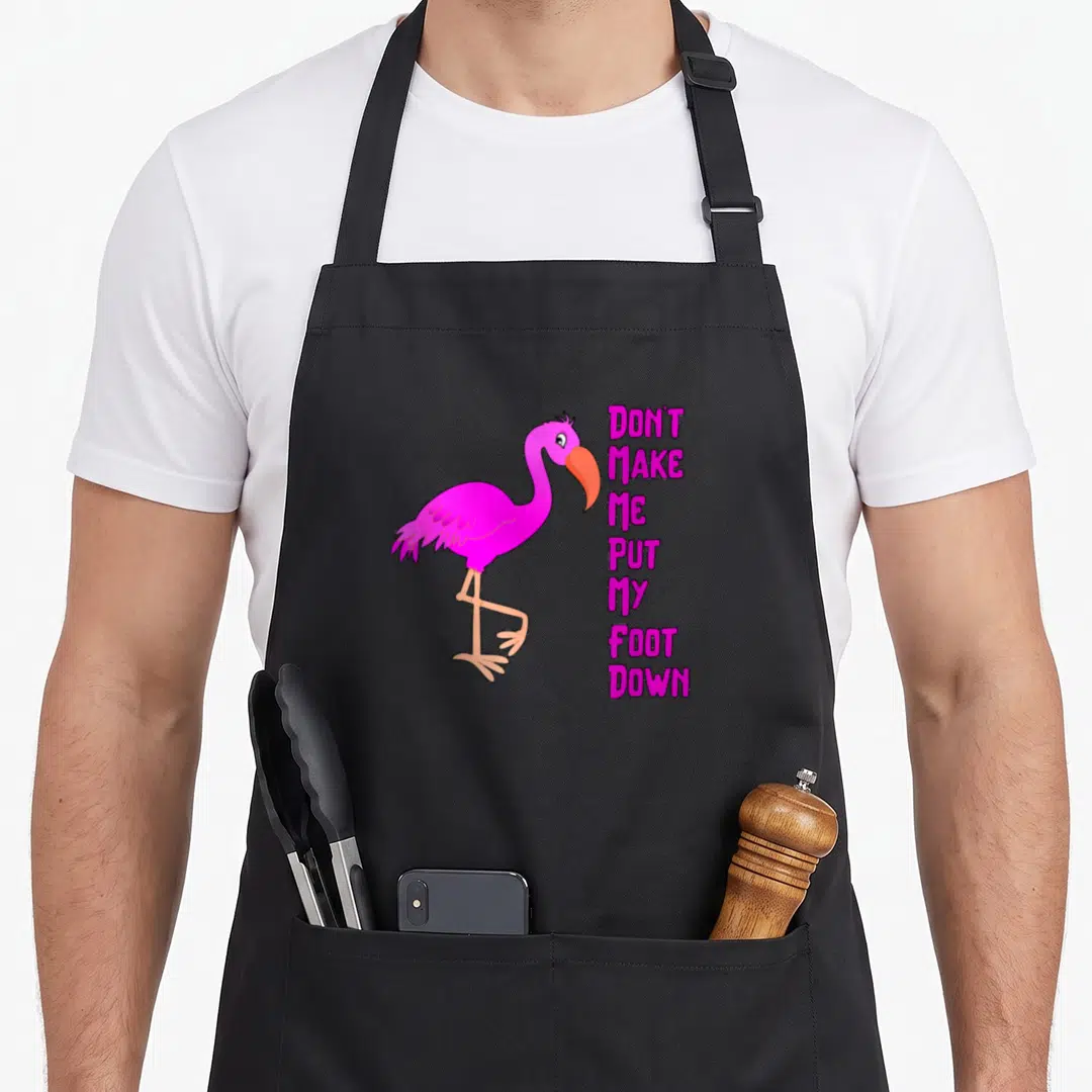 Retro awareness flamingo apron - Cartoon pink flamingo with orange beak next to stacked magenta text reading DON'T MAKE — shown on a black cotton canvas apron against a white background