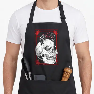 Japanese face apron - White horned skull in red geometric frame with small red flowers scattered around borders — shown on a black cotton canvas apron against a white background