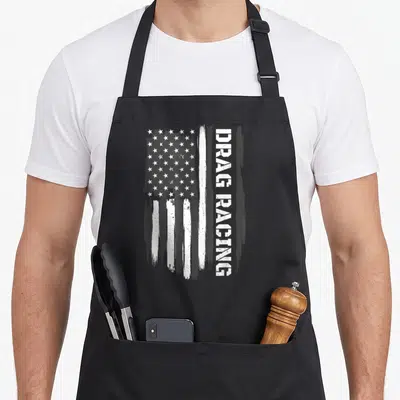 Race american flag apron - Distressed white USA flag next to vertical DRAG RACING text in monochrome — shown on a black cotton canvas apron against a white background