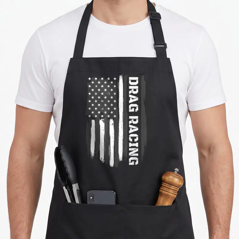 Race american flag apron - Distressed white USA flag next to vertical DRAG RACING text in monochrome — shown on a black cotton canvas apron against a white background