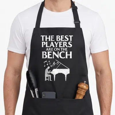 Keyboard player piano apron - Distressed white text and silhouette of person playing grand piano with floating musical — shown on a black cotton canvas apron against a white background