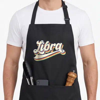 Zodiac september birthday apron - Retro script text Libra with cream orange blue and green shadows — shown on a black cotton canvas apron against a white background