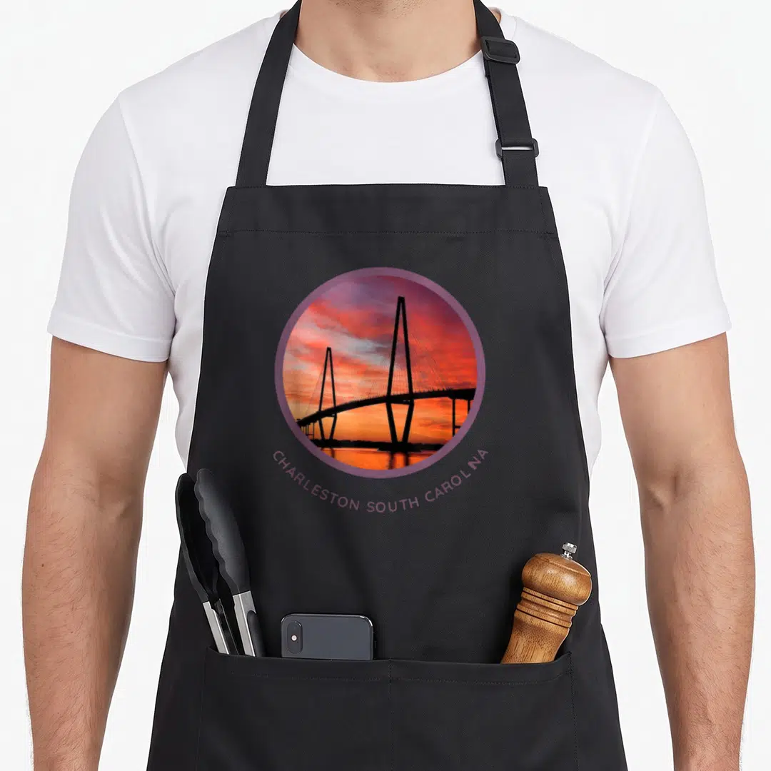 Ravenel charleston apron - Circular bridge sunset photo with curved Charleston South Carolina text in purple and orange — shown on a black cotton canvas apron against a white background