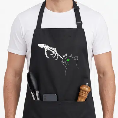 Boop skeleton apron - White skeletal hand pointing finger at a cat's nose with green eye and text BOOP — shown on a black cotton canvas apron against a white background