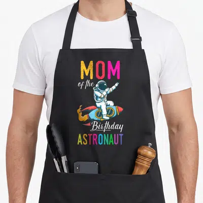 Space bday birthday apron - MOM of the Birthday ASTRONAUT text with cartoon astronaut on rocket in rainbow colors — shown on a black cotton canvas apron against a white background