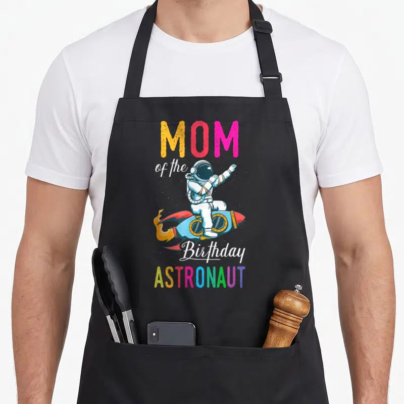 Space bday birthday apron - MOM of the Birthday ASTRONAUT text with cartoon astronaut on rocket in rainbow colors — shown on a black cotton canvas apron against a white background
