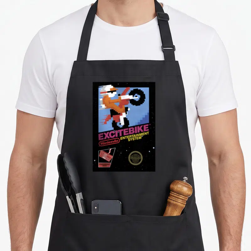 Excitebike game aprons - Pixelated motorcyclist, stacked title text, game cartridge icon, and gold seal on black starry — shown on a black cotton canvas apron against a white background
