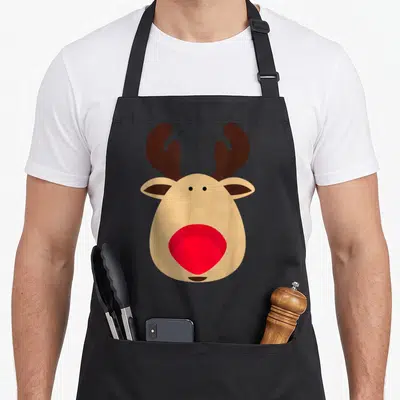 Reindeer red christmas apron - Cartoon reindeer head with red nose and brown antlers on tan background — shown on a black cotton canvas apron against a white background
