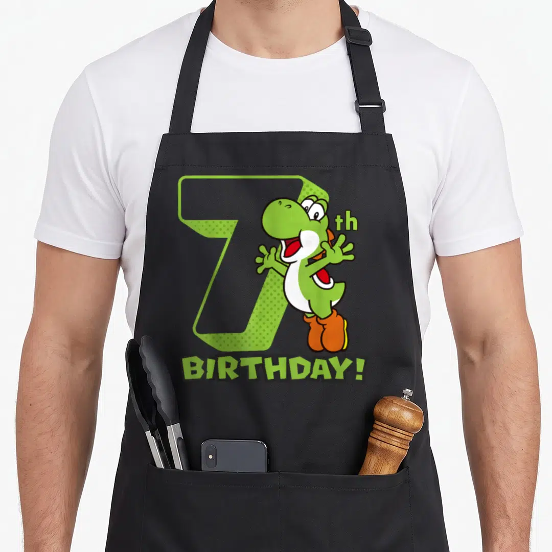 7th holiday apron - Green cartoon dinosaur with orange shoes standing in front of a large number 7 and birthday text — shown on a black cotton canvas apron against a white background