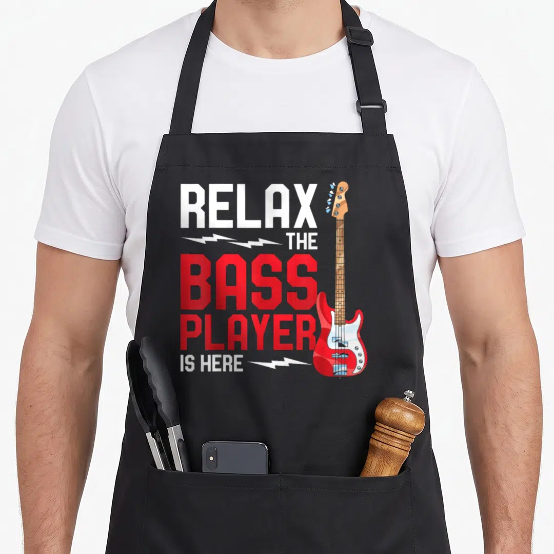 Entspann bassist apron - Red and white bass guitar with stacked text RELAX THE BASS PLAYER IS HERE and lightning bolts — shown on a black cotton canvas apron against a white background