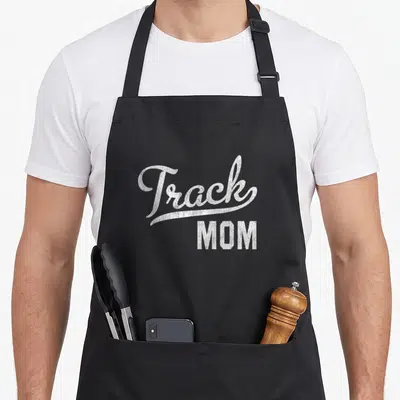 Proud track apron - White distressed Track MOM typography on black apron — shown on a black cotton canvas apron against a white background