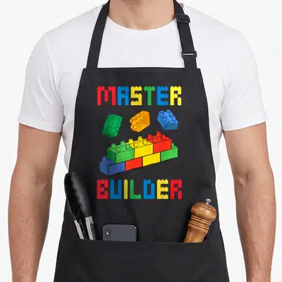 Block grill master apron - MASTER BUILDER text flanking red blue yellow and green building blocks in cartoon style — shown on a black cotton canvas apron against a white background