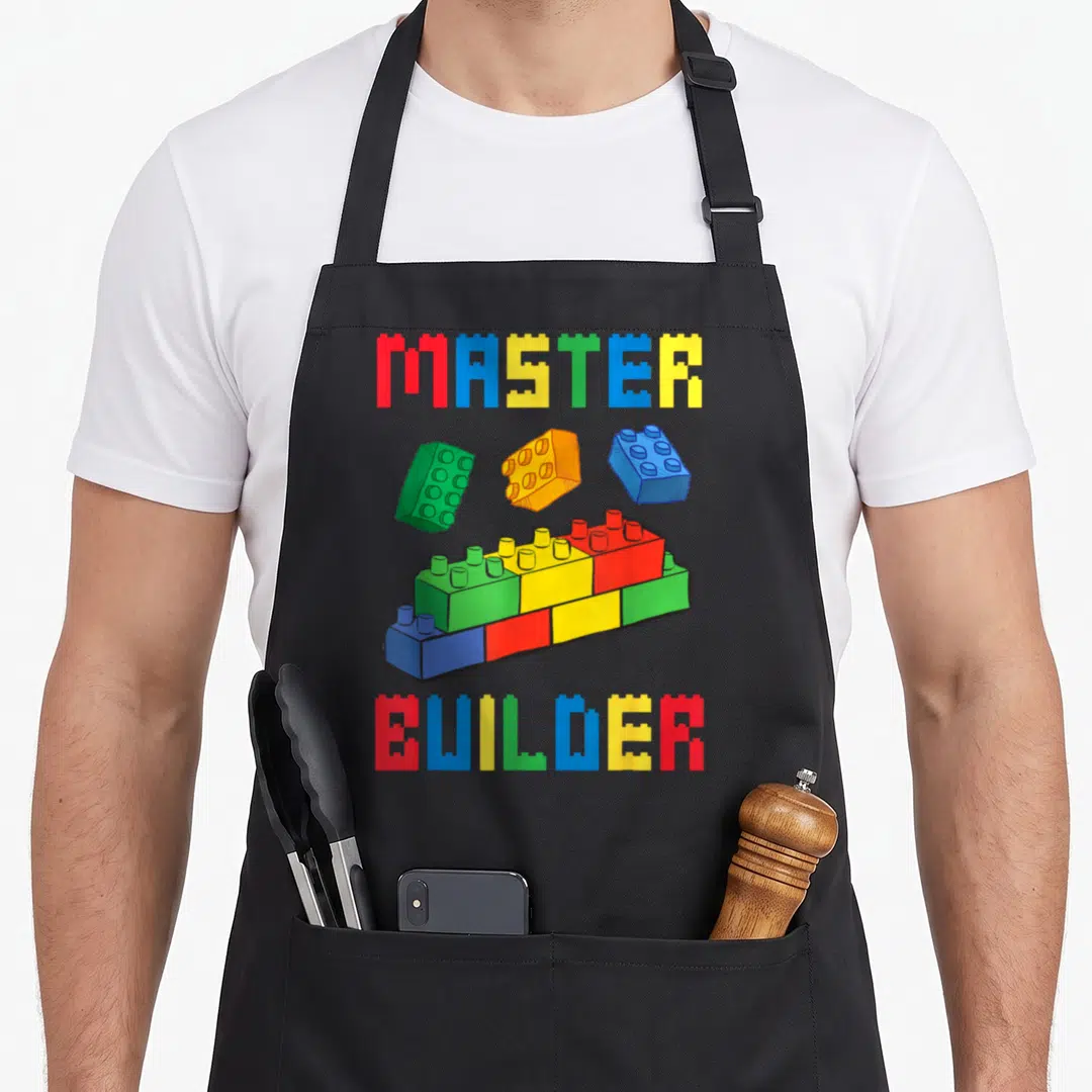Block grill master apron - MASTER BUILDER text flanking red blue yellow and green building blocks in cartoon style — shown on a black cotton canvas apron against a white background