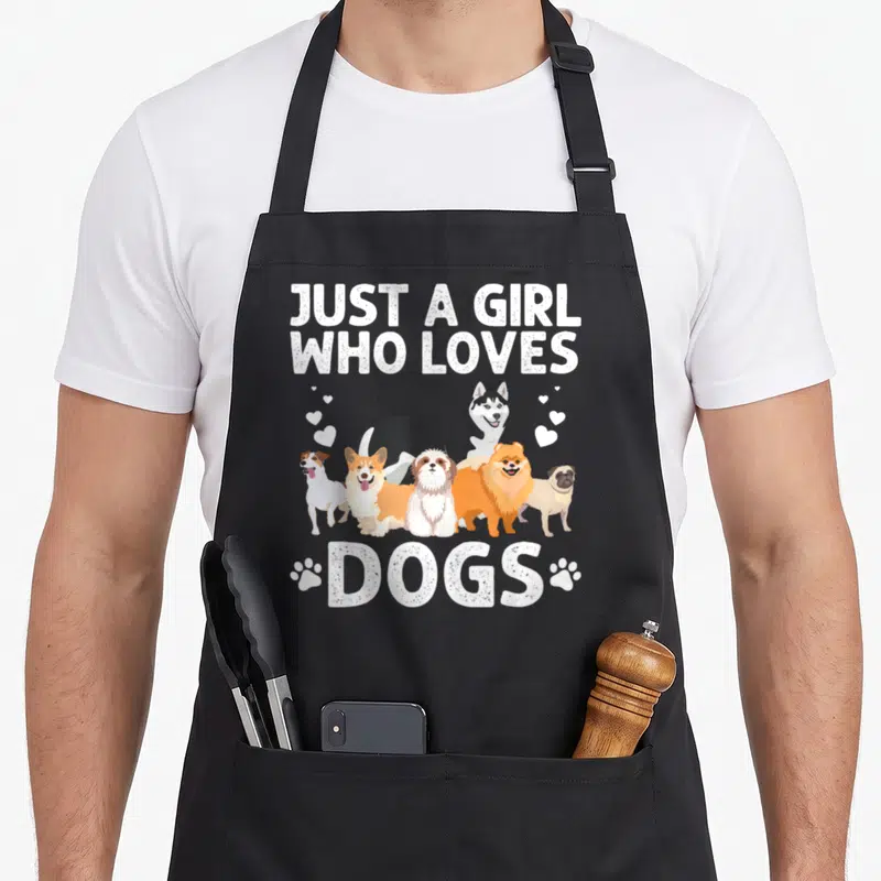 Handler dog groomer apron - Six cartoon dogs between Just a girl who loves dogs text and paw prints — shown on a black cotton canvas apron against a white background