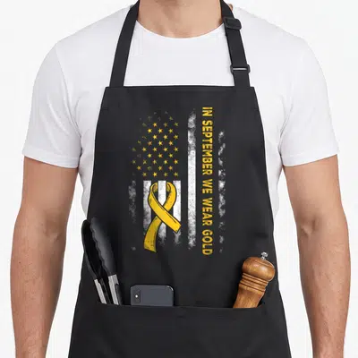 September us aprons - Yellow ribbon over white stripes with gold stars and vertical text reading IN SEPTEMBER WE WEAR — shown on a black cotton canvas apron against a white background