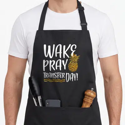 Wake infertility awareness apron - WAKE PRAY TRANSFER DAY! text with gold pineapple and wavy line — shown on a black cotton canvas apron against a white background