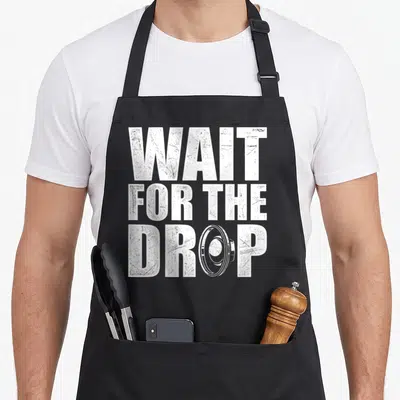 Wait dubstep apron - White stacked text WAIT FOR THE DROP with camera lens replacing the letter O — shown on a black cotton canvas apron against a white background
