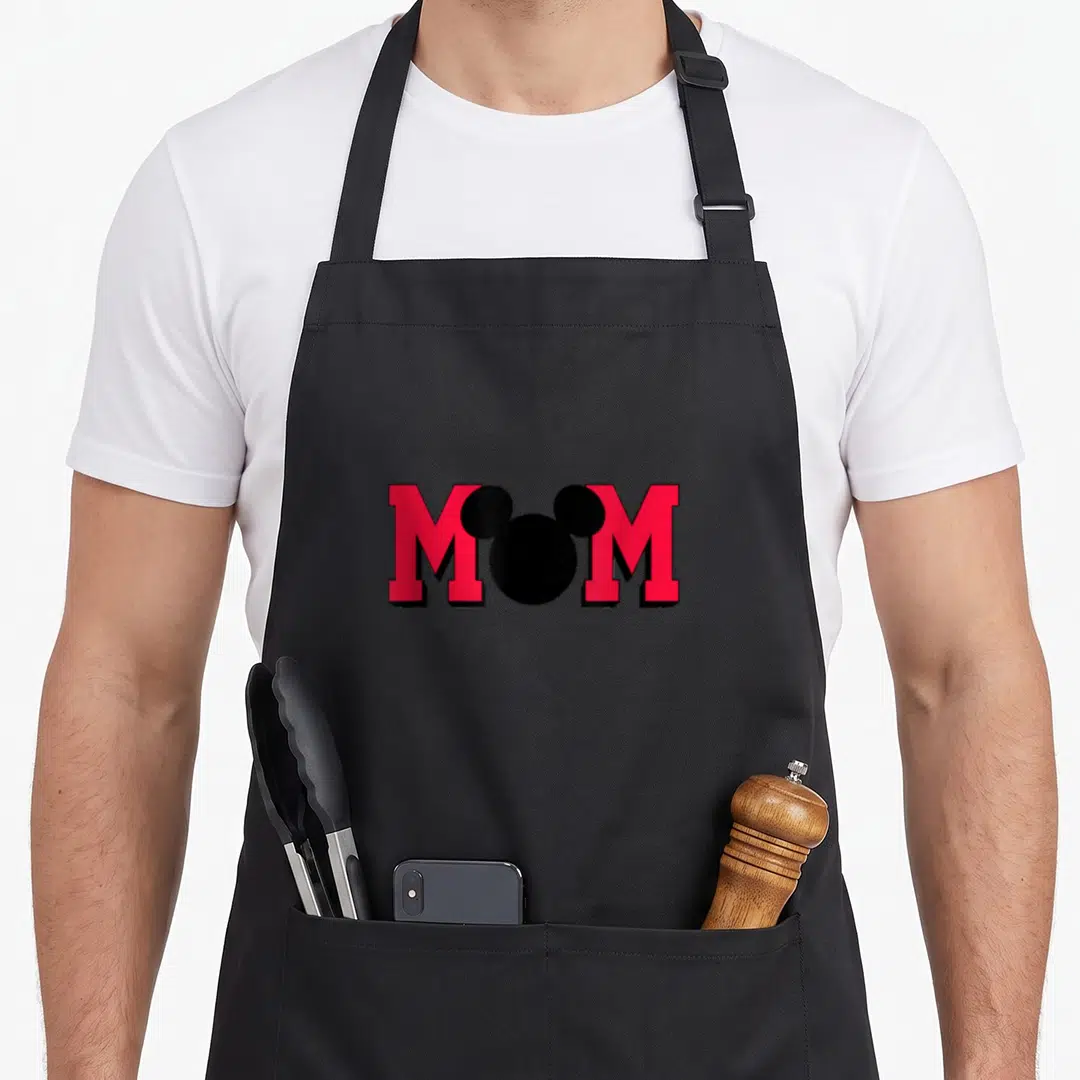 Silhouette mouse mom apron - MOM text in red with black Mickey head silhouette replacing the O — shown on a black cotton canvas apron against a white background