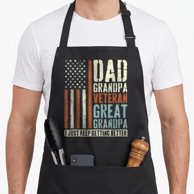 Vintage veteran grandpa apron - Distressed USA flag next to stacked words Dad Grandpa Veteran Great Grandpa I just keep — shown on a black cotton canvas apron against a white background