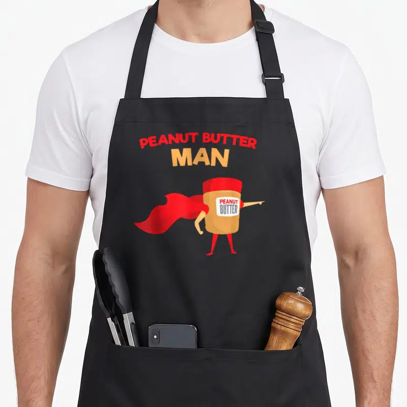 Superhero butter apron - Cartoon peanut butter jar man with red cape pointing right under stacked text — shown on a black cotton canvas apron against a white background