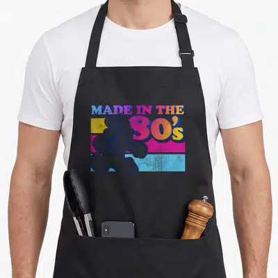 Gradient 80s apron - Mario character over blue pink yellow stripes with Made In The 80s text — shown on a black cotton canvas apron against a white background