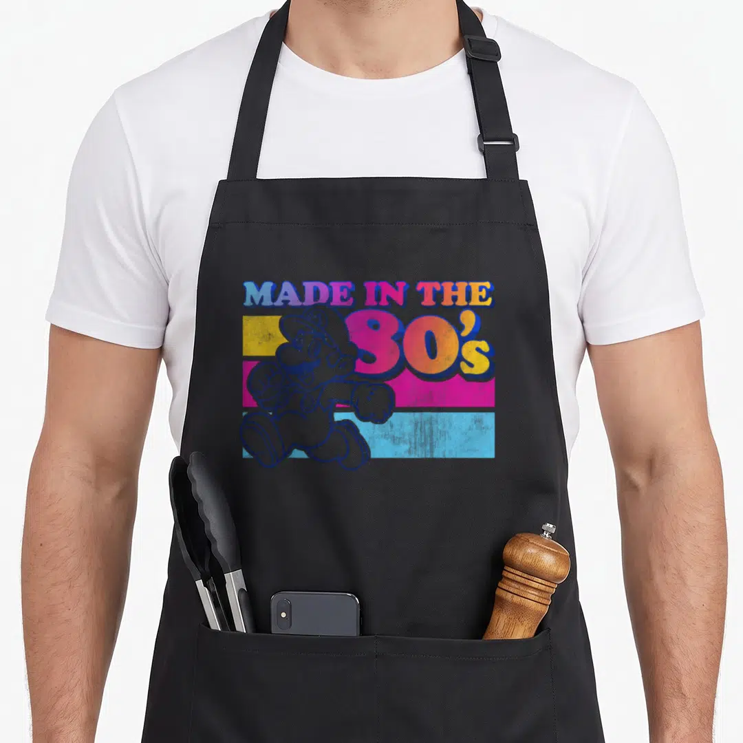 Gradient 80s apron - Mario character over blue pink yellow stripes with Made In The 80s text — shown on a black cotton canvas apron against a white background