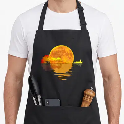 Moon acoustics apron - Minimalist moon in yellow orange and red reflecting over water with trees and city skyline — shown on a black cotton canvas apron against a white background