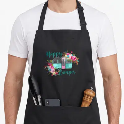 Vintage happy camper apron - Teal camper trailer with pink door surrounded by floral bouquet and teal script text — shown on a black cotton canvas apron against a white background