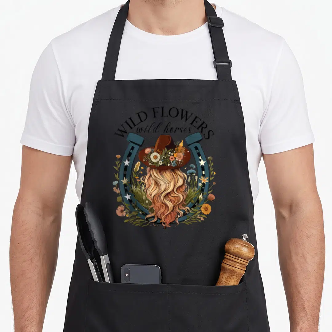 Wildflower country apron - Woman with blonde hair in floral hat inside blue-grey horseshoe with WILD FLOWERS text — shown on a black cotton canvas apron against a white background