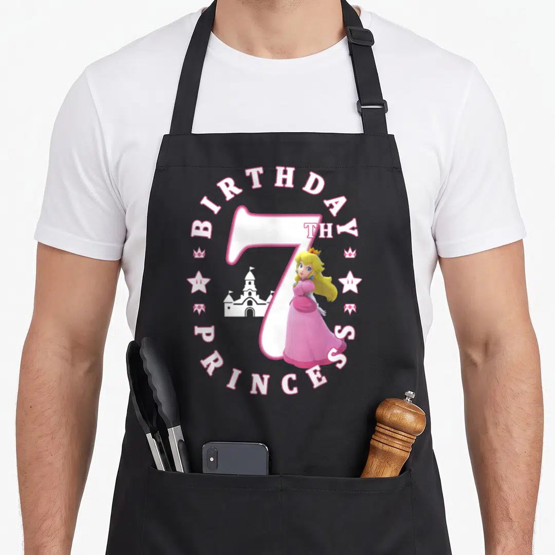 7th princess apron - Princess Peach stands before a number 7 surrounded by BIRTHDAY, TH, PRINCESS text, crowns — shown on a black cotton canvas apron against a white background