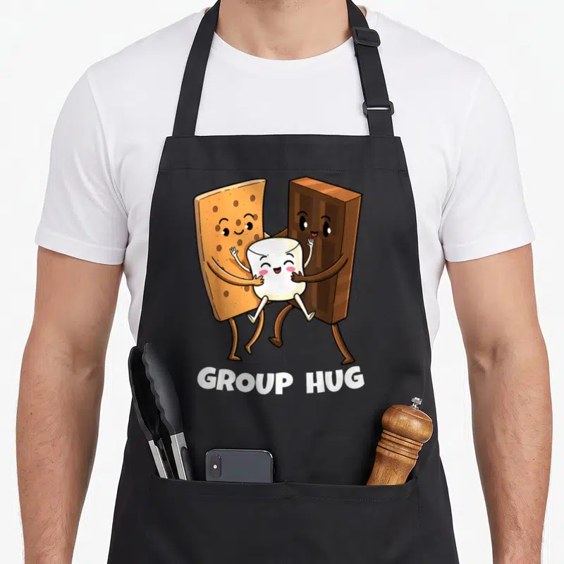 Smore marshmallow camping apron - Cartoon graham cracker and chocolate hugging marshmallow with GROUP HUG text — shown on a black cotton canvas apron against a white background