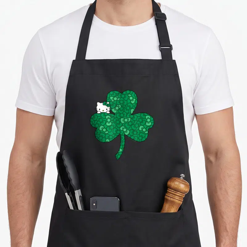 Clover bow apron - Green shamrock with bow pattern and white character peeking from behind top-left leaf — shown on a black cotton canvas apron against a white background