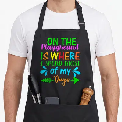 Funny spend playground apron - Stacked colorful text reading On The Playground Is Where I Spend Most Of My Days — shown on a black cotton canvas apron against a white background