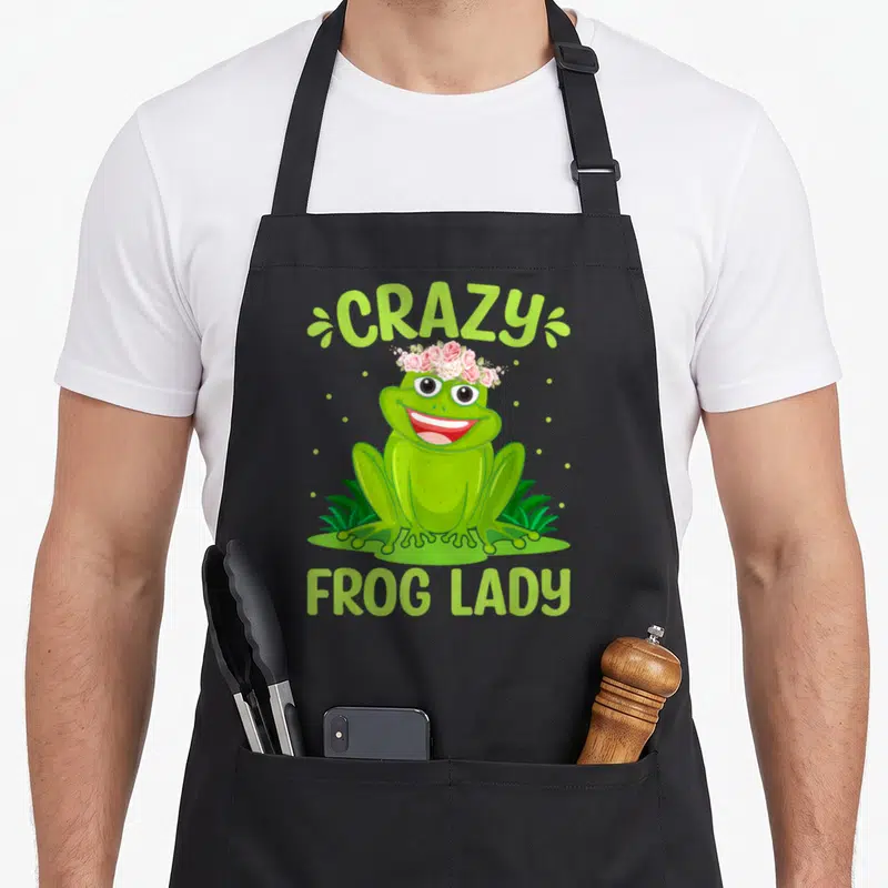 Funny frog floral apron - Cartoon green frog wearing pink flower crown with arched CRAZY and bottom FROG LADY text — shown on a black cotton canvas apron against a white background