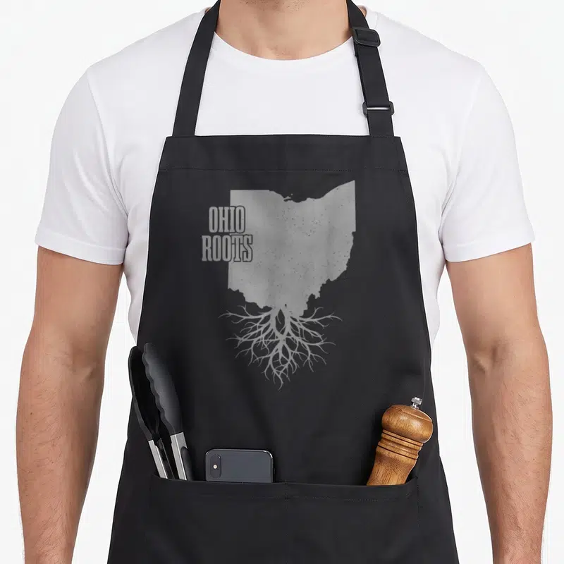 Root patriotic apron - Grey Ohio state map with tree roots and stacked OHIO ROOTS text — shown on a black cotton canvas apron against a white background