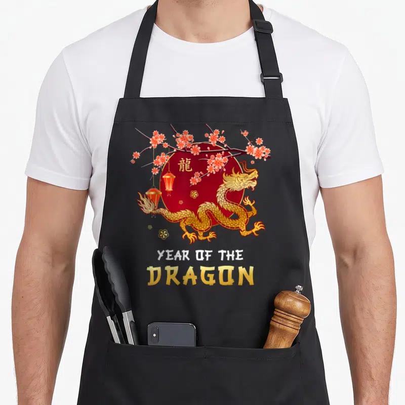 Lunar dragon apron - Gold dragon on red circle with pink blossoms, lanterns, and Chinese character — shown on a black cotton canvas apron against a white background
