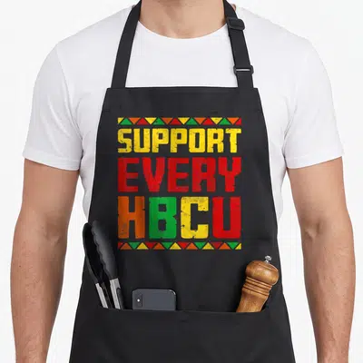 Historically graduate apron - Stacked colorful text reading SUPPORT EVERY HBCU with distressed finish and triangle — shown on a black cotton canvas apron against a white background