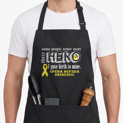 Warrior spina bifida awareness apron - Yellow ribbon and white text reading I gave birth to mine Hero with baby — shown on a black cotton canvas apron against a white background
