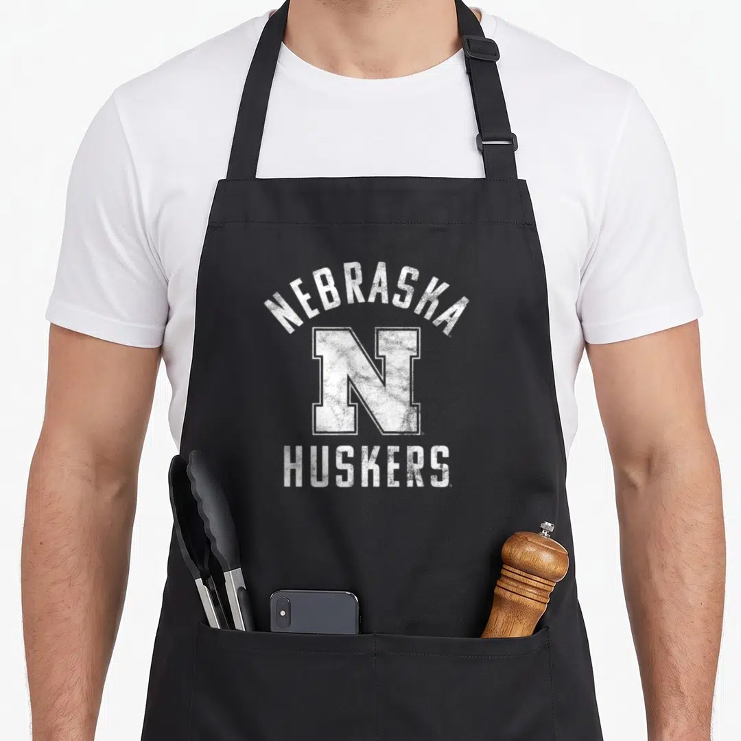 University nebraska girl apron - White distressed text reading NEBRASKA, N, and HUSKERS stacked vertically — shown on a black cotton canvas apron against a white background