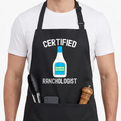 Ranch sauce dressing apron - Certified Ranchologist text over cartoon blue and green ranch dressing bottle — shown on a black cotton canvas apron against a white background