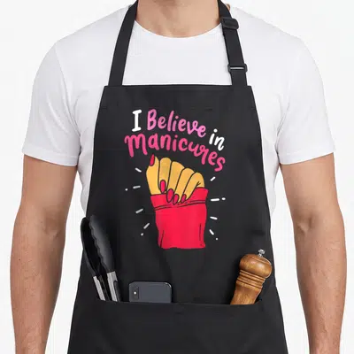 Beautiful manicurist apron - Arched I Believe in Manicures text above cartoon hand with red nails and white sparkles — shown on a black cotton canvas apron against a white background